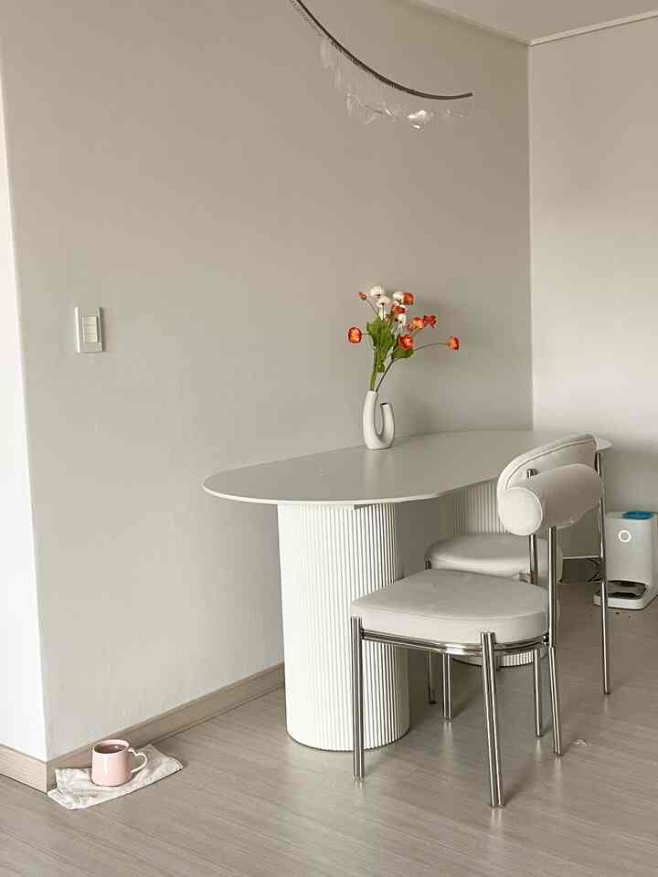 White-toned small kitchen space featuring a Mid-Century Modern dining table and chairs, vase with flowers, and a dog automatic feeder in a simple interior