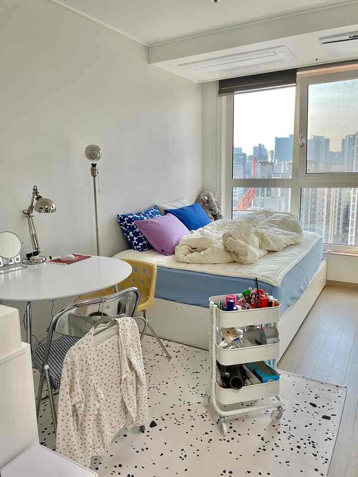 明るいホワイトとブルーの色調の縦長ワンルーム寝室、コンパクトなベッドと丸いダイニングテーブルが配置された雰囲気のある空間