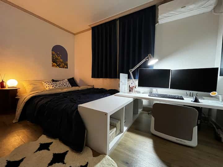 A modern and cozy studio apartment bedroom in black and white tones featuring a compact desk and blackout curtains