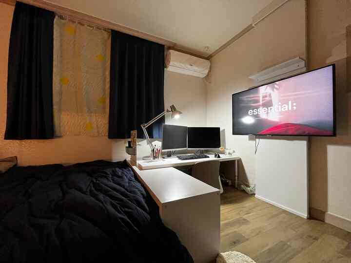 A small bedroom home office space in white and black tones, featuring a desk with dual monitors and a wall-mounted TV, creating a modern, focused atmosphere