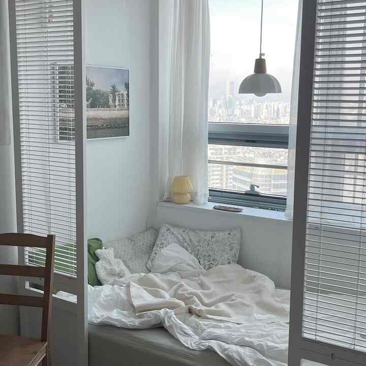 White-toned bedroom interior showcasing a bed with pillows and blankets, separated by blinds and room dividers, creating a simple and cozy atmosphere
