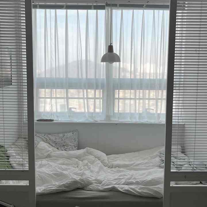 ホワイトトーンのワンルーム寝室、自然光の下で快適な寝具とブラインドが調和したシンプルなインテリア