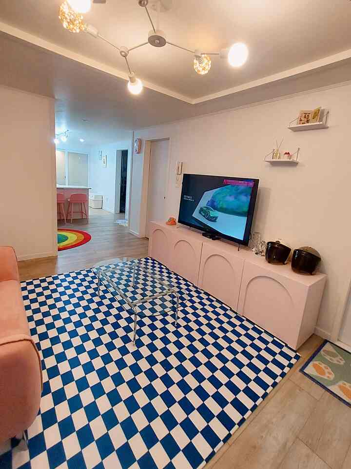 ピンクの家具と透明なガラステーブル、青のチェック柄ラグが配されたリビングルームと、その奥に見えるキッチンが調和した空間