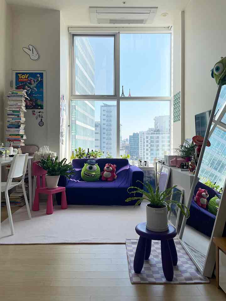 Bright natural light fills a compact single household living room featuring a purple sofa, plants, full-length mirror, and workspace desk