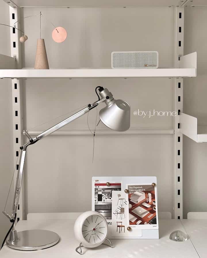 Minimal desk area in white and silver tones featuring a clock, table lamp, and wooden mobile with clean aesthetics