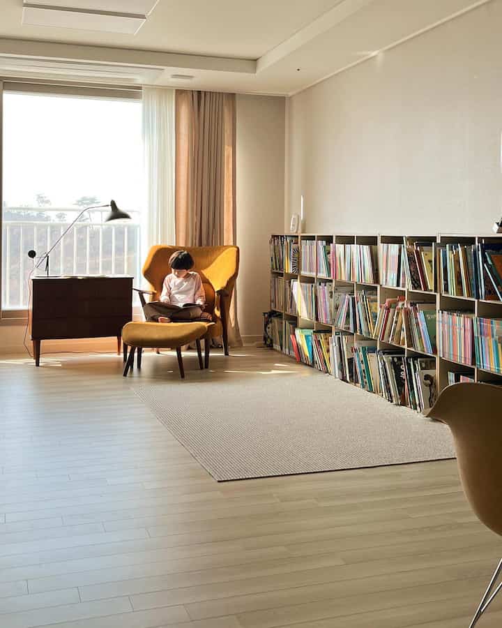 Natural-toned living room with abundant natural light featuring an armchair where a child reads, alongside a large bookshelf creating a cozy atmosphere