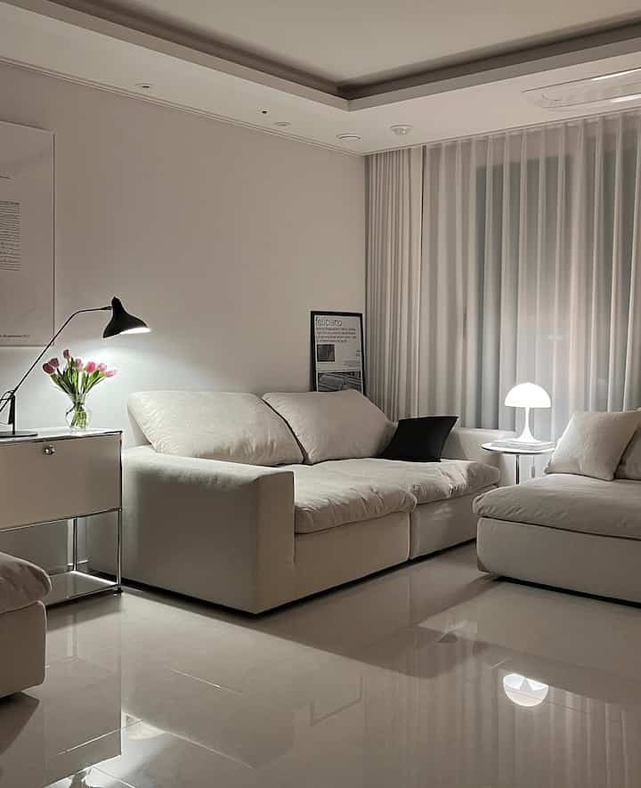 Bright white-toned living room featuring spacious fabric sofas, curtains, modern table lamps, and a poster creating a cozy atmosphere