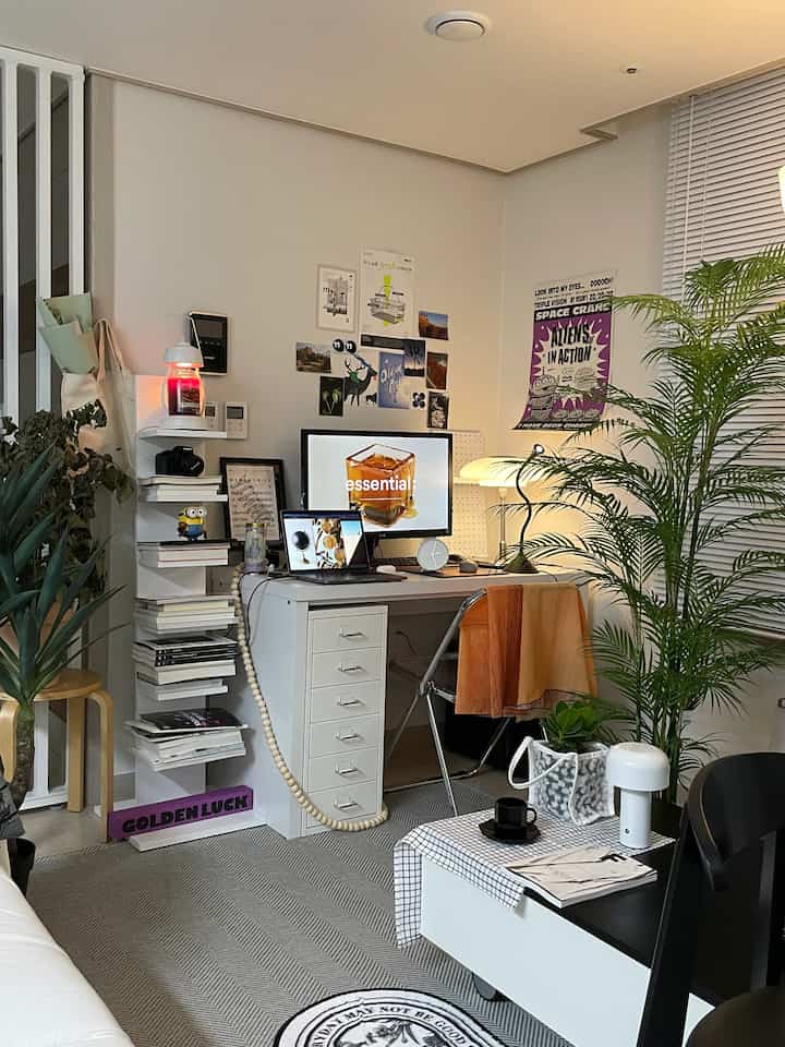 Cool-toned studio living room and home office featuring white desk, transparent chair, monitors, various lighting, and artificial plants creating a cozy atmosphere