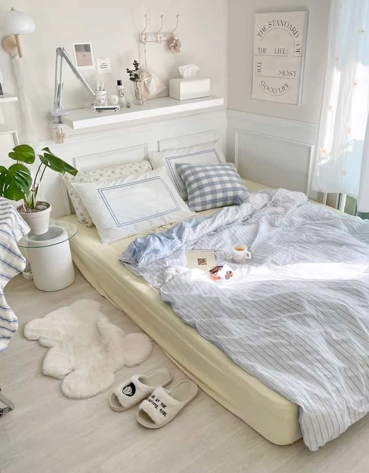 White and blue toned studio bedroom featuring clean bedding, cushions, and a teddy bear-shaped rug creating a cozy, bright atmosphere