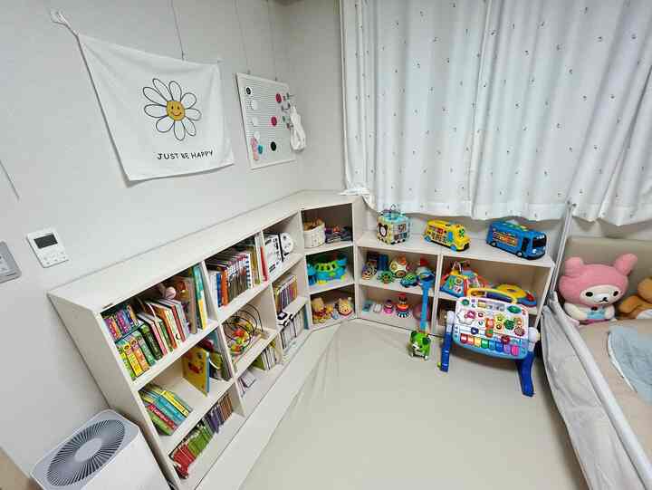 白を基調とした赤ちゃんの部屋、コーナー棚や本棚に本とおもちゃが整然と配置され、ベビーベッドとカーテンがある温かみのあるインテリア