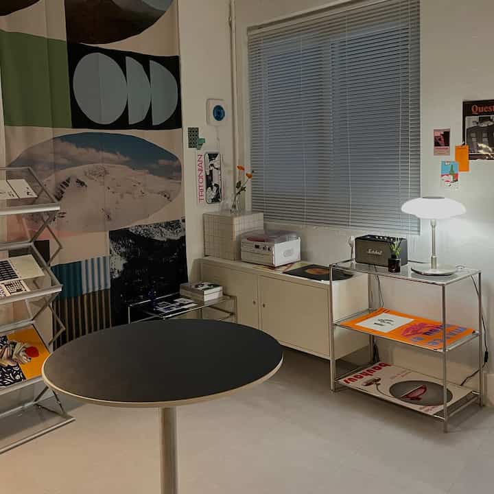 ホワイトとベージュのトーンのモダンなリビングルーム、アルミブラインドとメタルシェルフ、丸テーブルが配置された印象的な空間