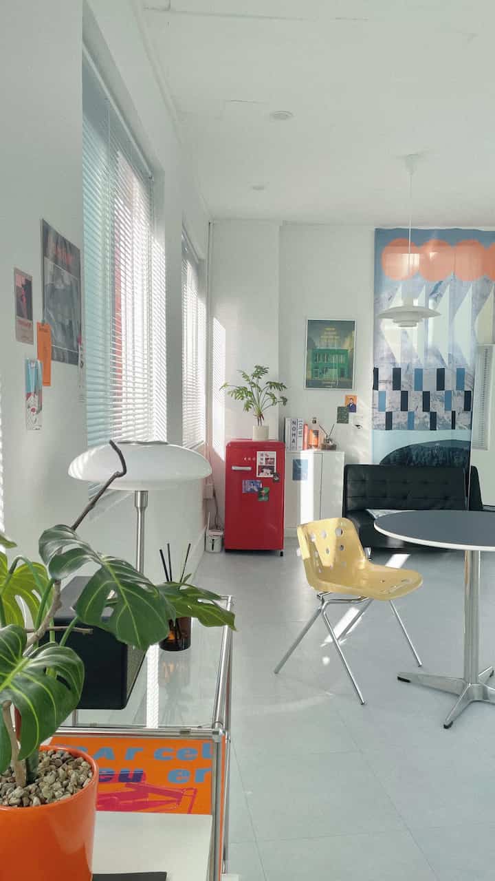 A white-tone studio living and kitchen space featuring a red mini refrigerator, yellow dining chair, and mid-century modern style elements