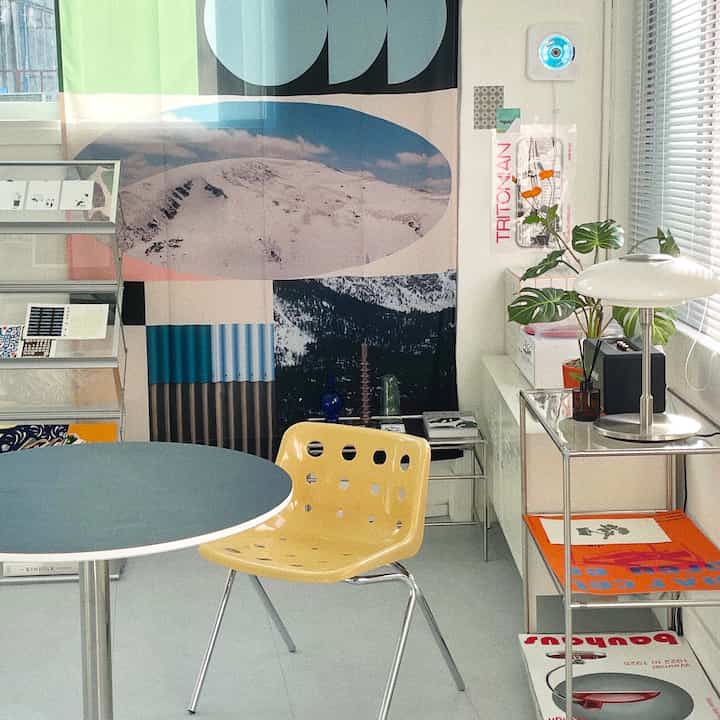 White-walled living space with blue table and yellow chair in Mid-Century Modern style, featuring large mountain poster and plants