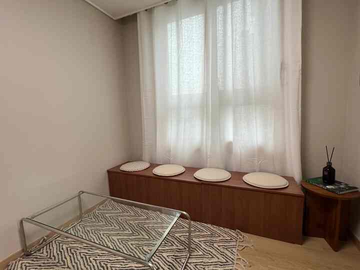 ナチュラルな色調のリビング、木目調のベンチと白いシアーカーテンが配置された雰囲気のある空間