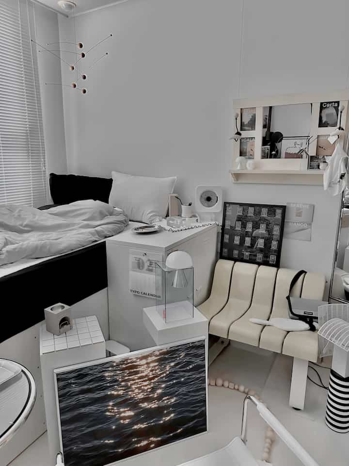 モノトーンの小さな寝室、白い寝具と黒いベッドフレーム、ミッドセンチュリーモダンのベンチと額縁が配置された整った空間