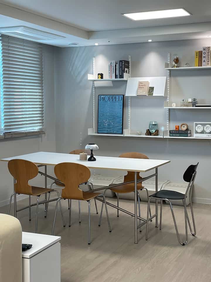 A dining room featuring white and wood tones, Mid-Century Modern dining chairs, wall shelves, and posters arranged neatly