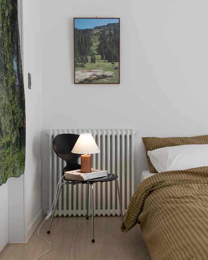 A bedroom with white walls and beige bedding, featuring a vintage black Ant chair and table lamp centrally placed in a simple setting