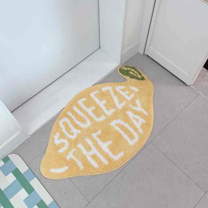 黄色のマンゴー形バスマットがグレーの床と白いドアの近くに配置された、爽やかな雰囲気の玄関スペース