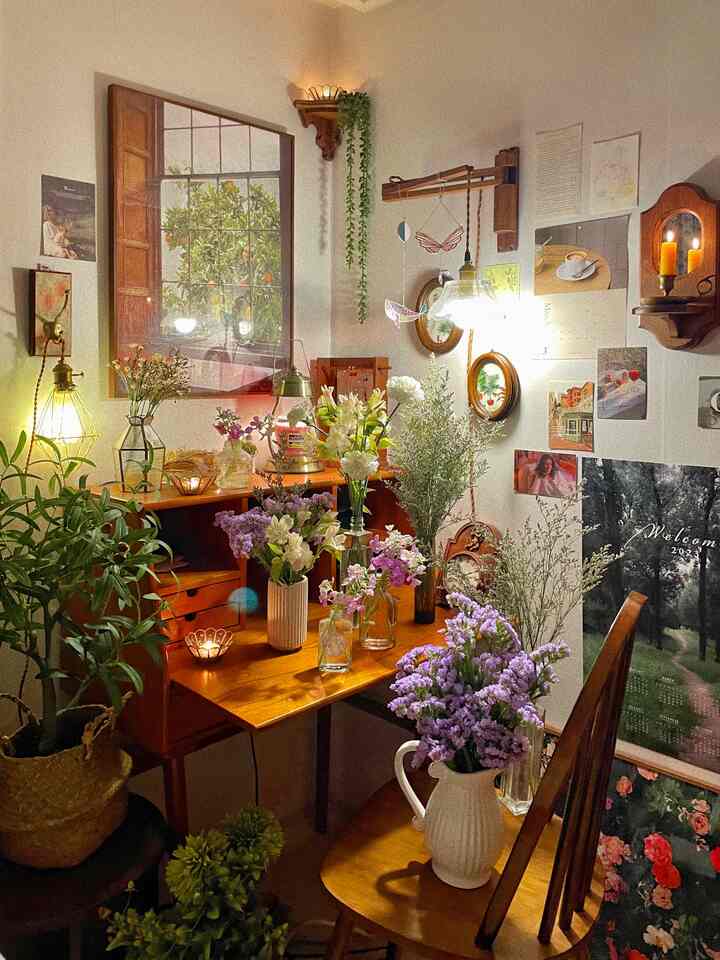 Cozy bedroom corner with vintage wooden desk and chair adorned with various flowers and candles, walls decorated with posters and frames