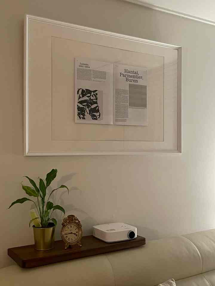 Natural color living room with beige sofa, a wooden shelf holding a gold clock and plant, and a framed picture on the wall