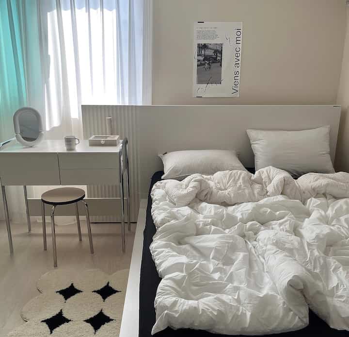 White and black toned bedroom featuring a tidy bed, vanity desk, and metal-legged stool in a simple modern setting