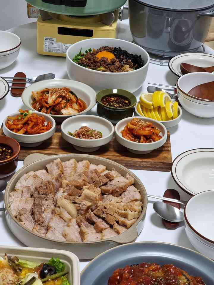 白いテーブルの上に韓国料理の食卓、ボッサムやビビンバなど多彩な器とおかずが配置された雰囲気のある空間