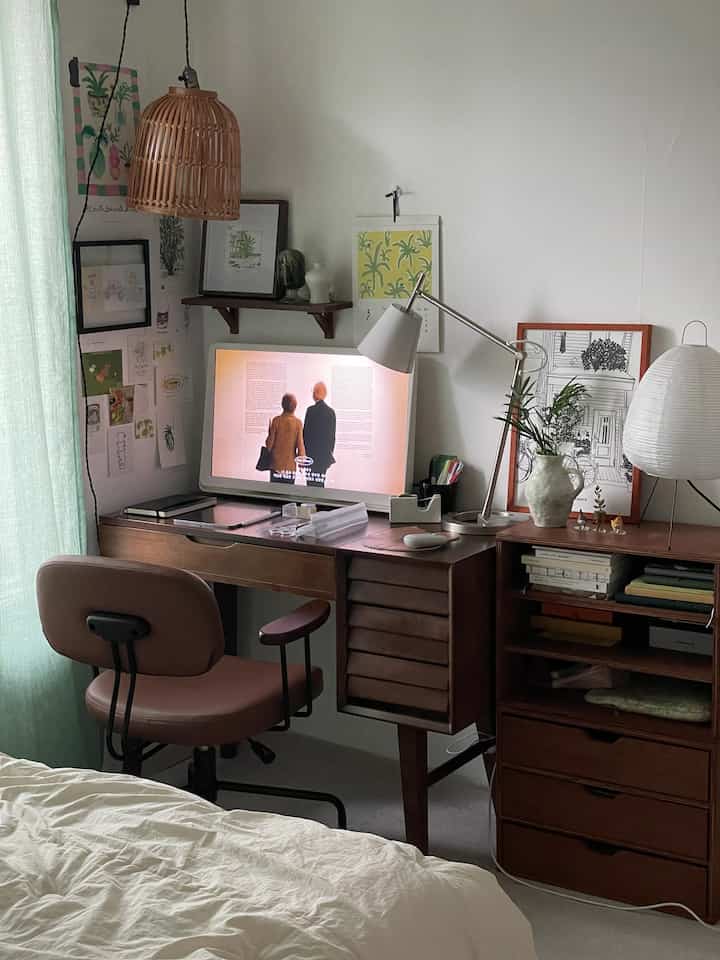 White and wood-tone bedroom combined with home office, featuring a wood desk, chair, computer, picture frames, and lamp creating a cozy atmosphere