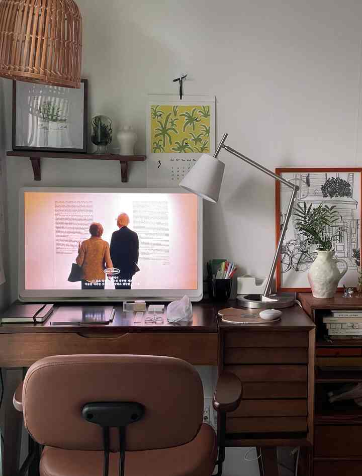 Modern home office with white walls and wood-tone furniture, featuring a desk with monitor and table lamp, creating a cozy atmosphere