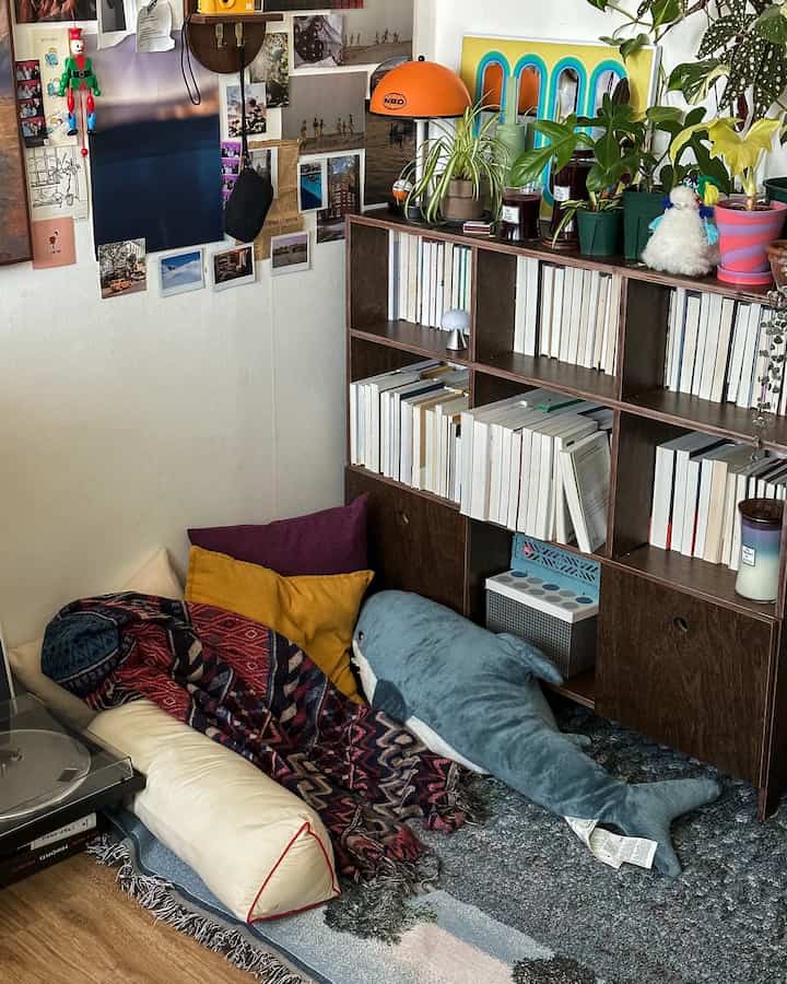 白い壁と木目調の本棚が配置されたリビングの一角、クッションと大型のサメのぬいぐるみがあり、居心地の良い自然な雰囲気のインテリア