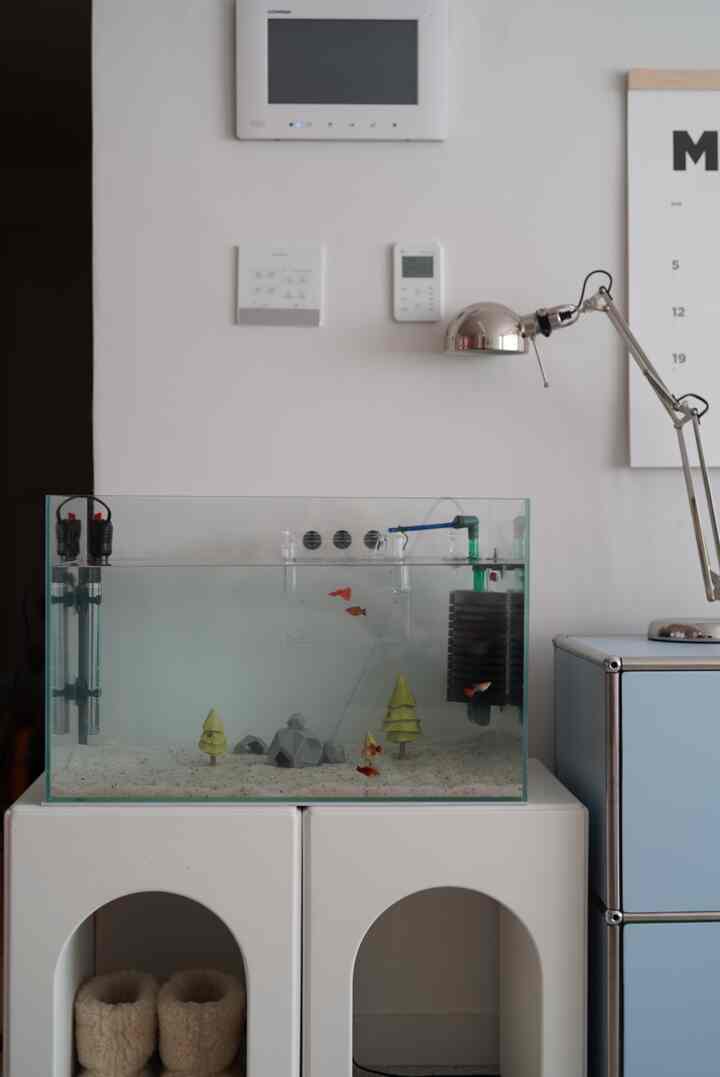 White and blue toned living room featuring a central aquarium with fish, boots on the bottom left, and a metal desk lamp on the right, creating a clean atmosphere