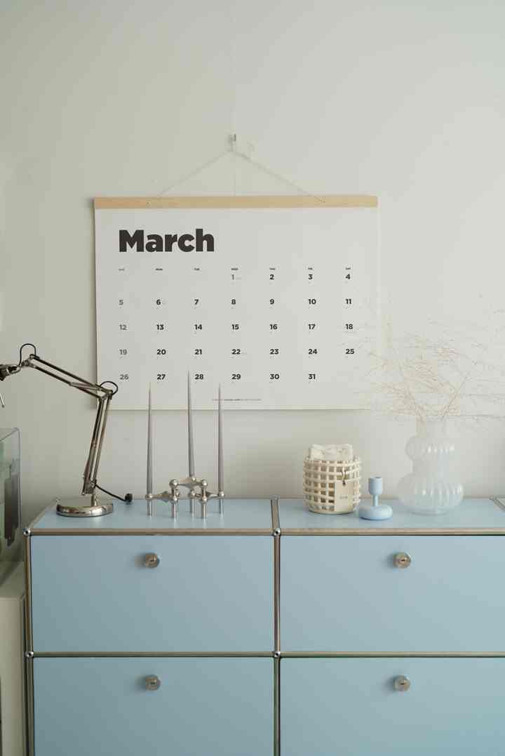 Minimal and clean space with white walls and pastel blue modular storage cabinet, featuring a March calendar on wall and candle holders with decor on cabinet top