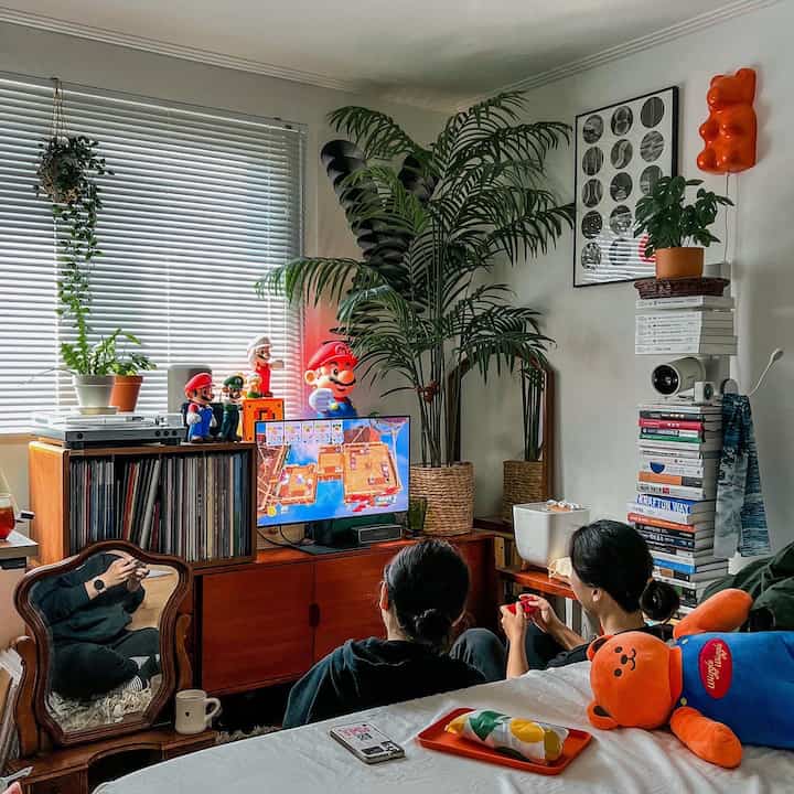 White-walled living and dining room with wood tone furniture, featuring Nintendo gaming setup and plants in a cozy home party space