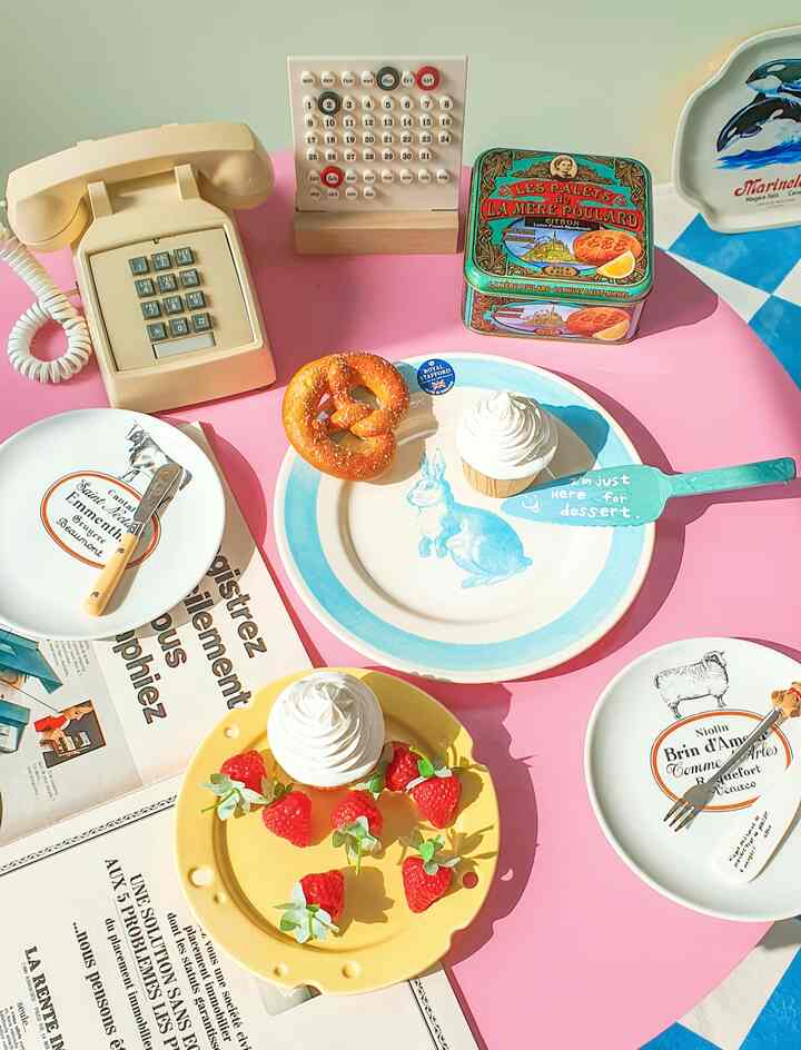 Cozy home cafe setting with a pink tablecloth-covered table featuring a retro phone, perpetual calendar, plates, and desserts