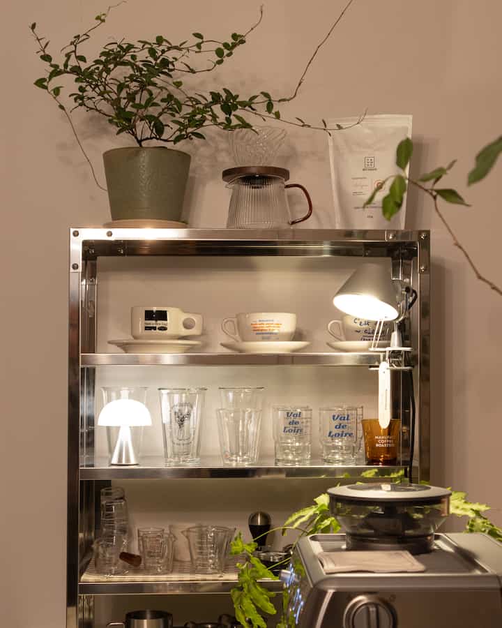 Cozy home cafe space with silver metal shelves, various coffee accessories, lighting, and green plants