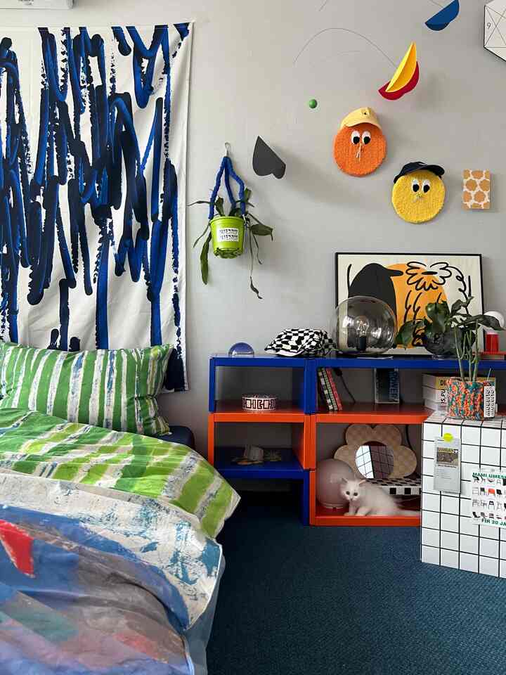 Beige-walled living room with blue carpet, featuring green patterned bedding and modern blue-orange shelving units