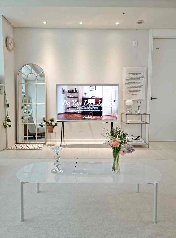 Bright white toned living room featuring a glass coffee table with vase and flowers, arched full-length mirror and textile poster on walls, modern ambiance