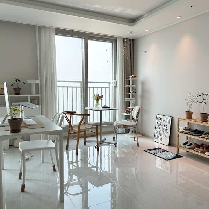 白とナチュラルな色調のリビング、窓辺に丸テーブルと椅子、左側に机とスツールが配置された清潔感のある明るい空間