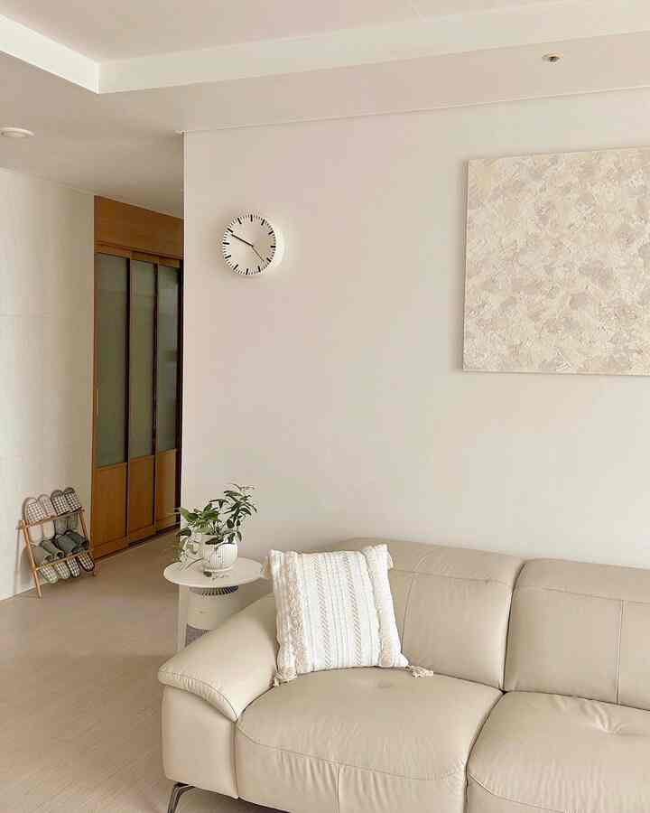 A white and beige toned living room featuring a leather sofa, cushion, wall clock, and textured wall art creating a cozy atmosphere.