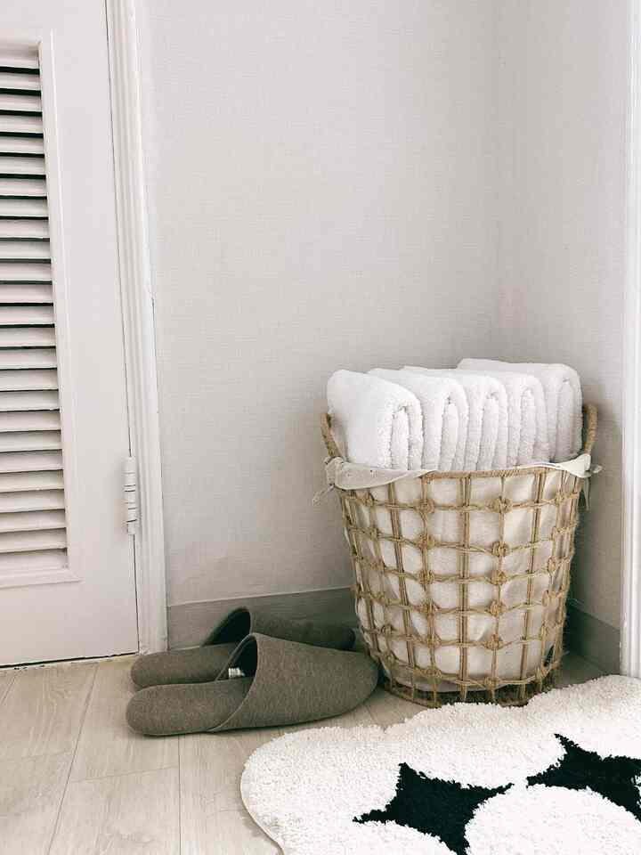 ナチュラルトーンのバスルームコーナー、ロープバスケットに並べられた白いタオル、ブラウンのスリッパ、白と黒のパターンラグが配置された温かみのある空間