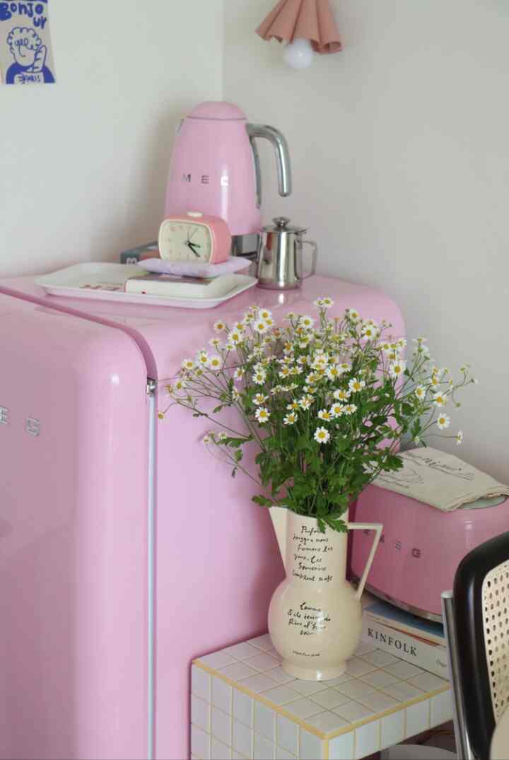 Cozy retro kitchen space in soft pink and white tones featuring a compact pink refrigerator and a vase of flowers