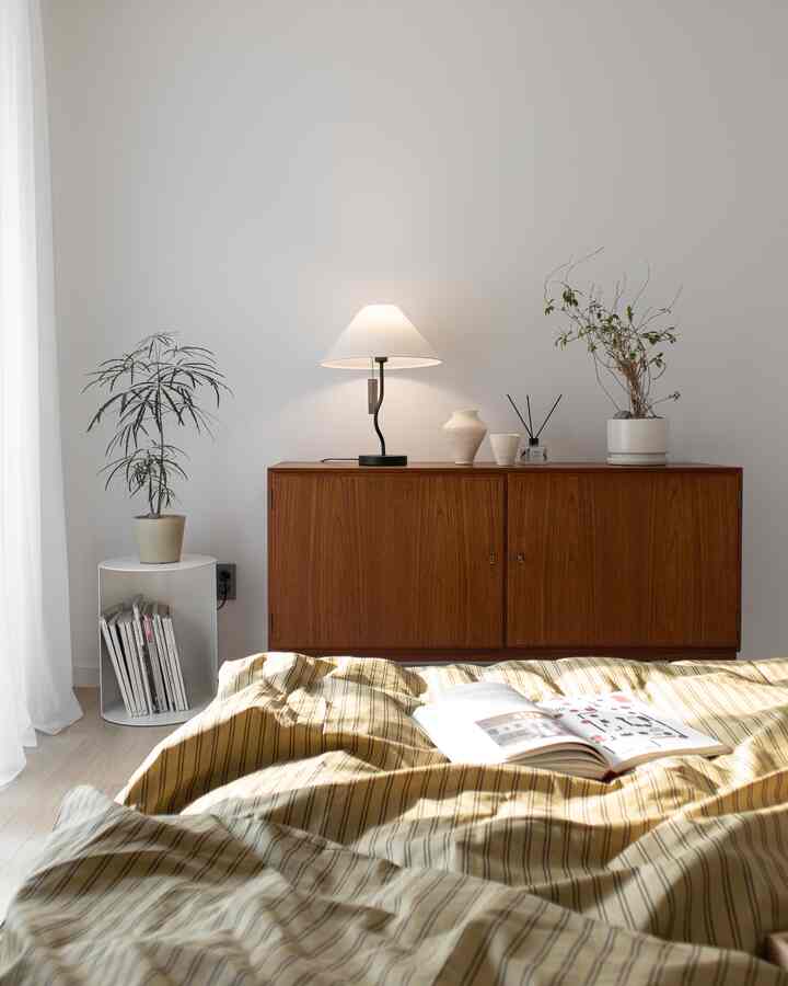 Natural bedroom with white walls, wood tone cabinet, plants, and a table lamp creating a cozy atmosphere