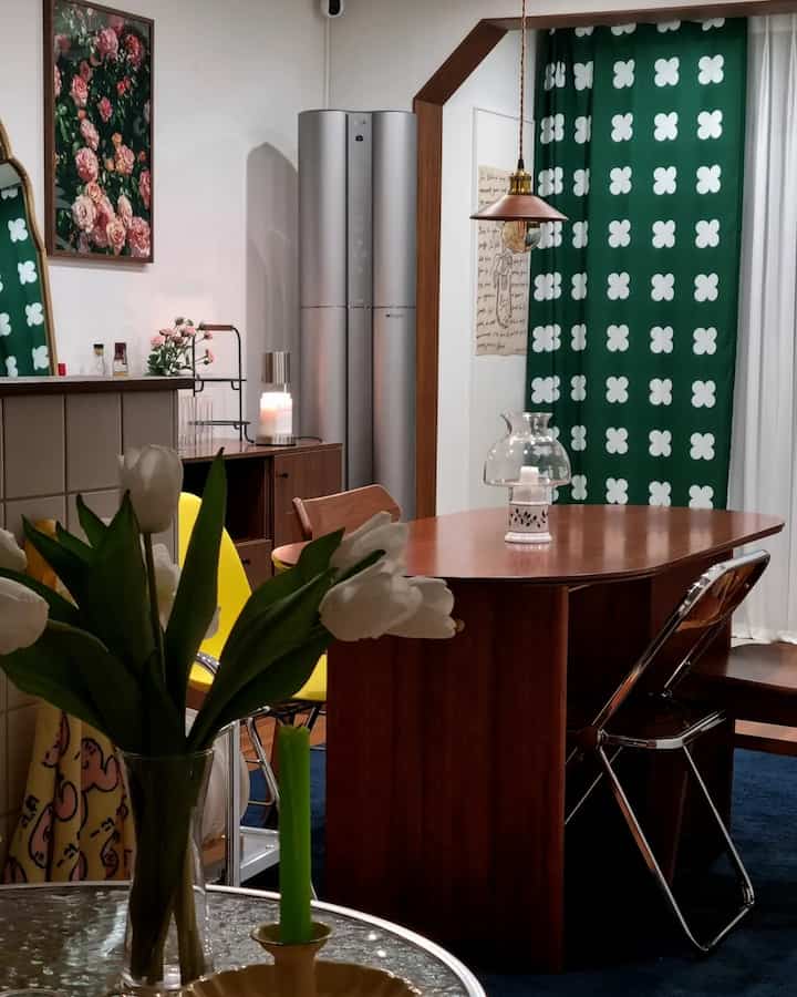 A wood-toned and green living and dining room featuring a wooden dining table, yellow chairs, and patterned curtains creating a cozy atmosphere