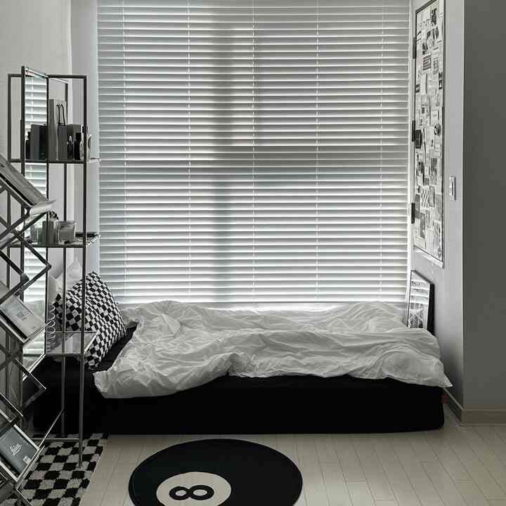 A modern bedroom with white and black tones, featuring blinds in the background, a bed, and a round 8-ball rug.