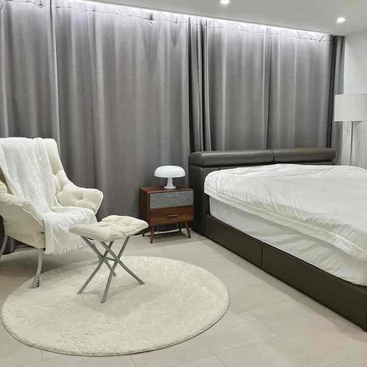 グレーのカーテンと白いラグがあるモダンな寝室、ダークブラウンのベッドとクリーム色のアームチェアが配置された落ち着いた読書コーナー