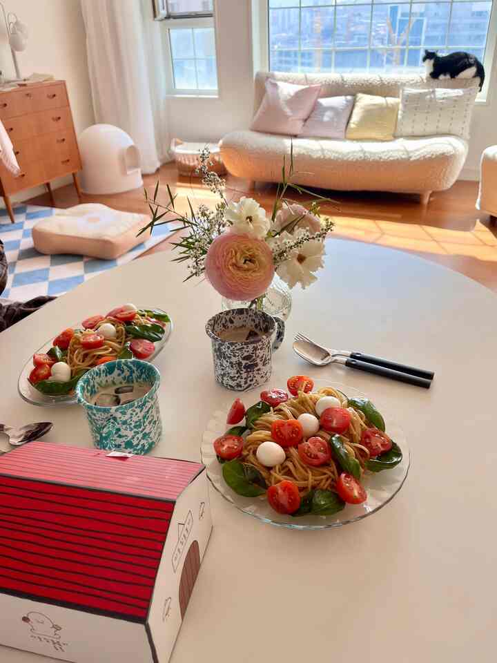 ベージュとホワイトのトーンが調和したリビング、中央のテーブルには花瓶とパスタが置かれ、窓辺に猫が座る温かみのあるインテリア