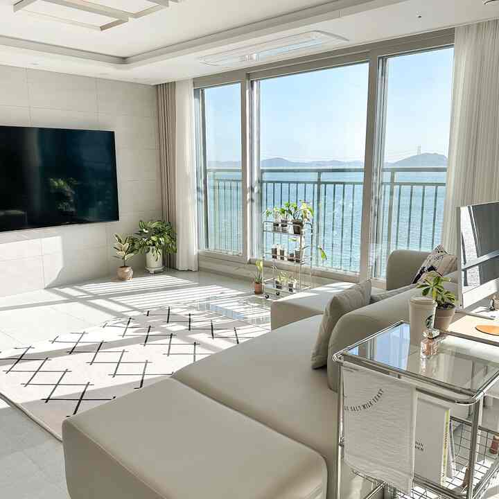 Bright modern living room in white and natural tones, featuring large windows with plants and sunlight filling the space