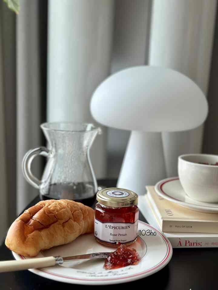 Black and white toned living space featuring a plate with croissant and rose jam, coffee carafe, and a table lamp creating a clean home cafe atmosphere