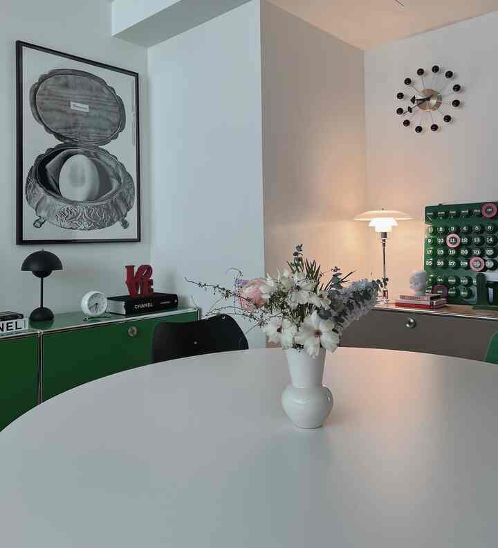 ホワイトトーンの壁とグリーンのサイドボードが配されたシンプルモダンなダイニングルーム、中央に花瓶が置かれた丸テーブルと多様な照明が調和する空間