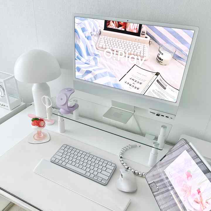 White-toned home office space featuring an iMac, keyboard, and glass monitor stand with a clean and modern workspace vibe
