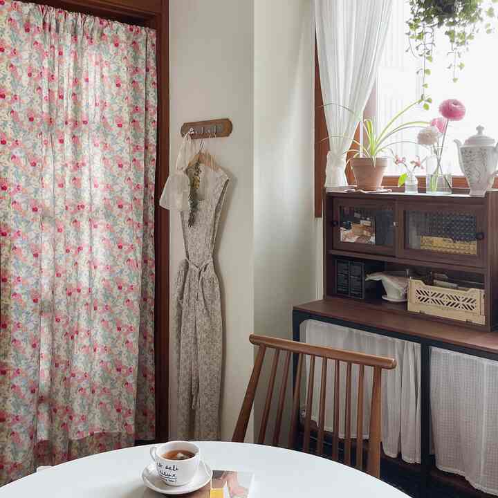 Natural and white-toned kitchen featuring floral curtain, wooden dining chair, and white eyelet curtain creating cozy atmosphere